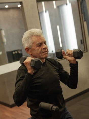 Un hombre de cabello canoso y camiseta negra sostiene dos mancuernas negras a la altura de sus hombros dentro de un gimnasio