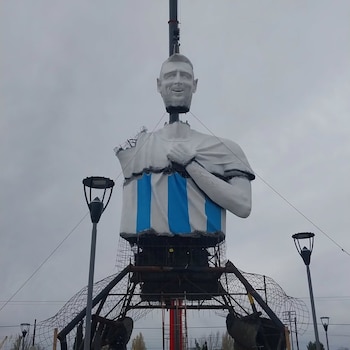 Vista de una enorme estatua de Lionel Messi vestido con la camiseta argentina, con el brazo levantado, frente a una multitud de personas con banderas