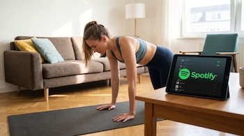 Mujer con cola de caballo en ropa deportiva haciendo ejercicio en una esterilla de yoga en casa, con una tablet mostrando el logo de Spotify sobre una mesa de madera.