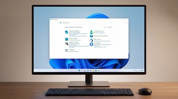 Un monitor negro muestra el Panel de Control de Windows 11. Un teclado y un ratón oscuros descansan sobre un escritorio de madera clara.