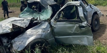 Una camioneta plateada con daños graves en la carrocería y el parabrisas destrozado se encuentra sobre el pasto junto a bomberos