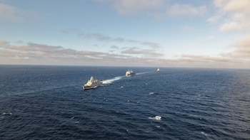 Tres buques navales de guerra de color gris navegan en fila sobre un océano azul bajo un cielo con nubes dispersas, dejando estelas blancas de espuma