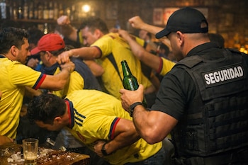 Grupo de hombres forcejea en un bar. Guardia de seguridad con uniforme negro y chaleco 'SEGURIDAD' sujeta botella verde. Hombres visten camisetas amarillas. Vaso con hielo en mesa.