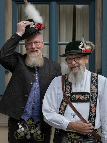 Dos hombres de barba blanca, gafas y sombreros con plumas, rosas y pines. Uno viste chaqueta y chaleco, el otro camisa blanca y tirantes bordados