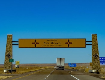 Un cartel vial con la inscripción Welcome to New Mexico sostenido por dos pilares de piedra sobre una carretera por la que avanza un camión