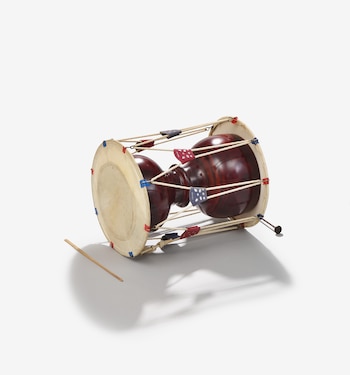 Un tambor janggu coreano de madera oscura y parches de piel clara yace de lado, con cuerdas tensadas y parches de tela colorida, junto a un baqueta de madera sobre un fondo blanco