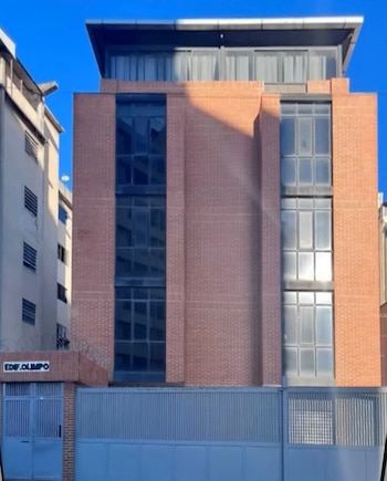 Vista frontal del Edificio Olimpo, un edificio de ladrillo de varios pisos con grandes ventanas oscuras y un techo moderno, detrás de una valla azul