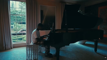 Hombre sentado tocando un piano de cola negro en una habitación. Se observa una ventana grande, cortinas, árboles y un coche en el exterior