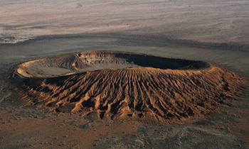 Vista aérea de un cráter volcánico con bordes dentados y un fondo oscuro, ubicado en un paisaje desértico plano de tonos marrones y ocres