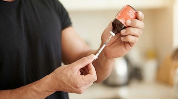 Manos de un hombre musculoso con camiseta negra extrayendo líquido de un frasco de vidrio ámbar con etiqueta naranja usando una jeringa médica. El fondo es borroso y claro.