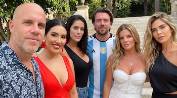Un hombre calvo, una mujer de rojo, una mujer de negro, un hombre con camiseta de Argentina, y dos mujeres rubias posan sonriendo al aire libre.