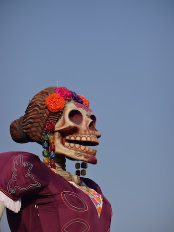 Estructura de una calavera con peinado trenzado, flores de colores, aretes largos y vestido morado sobre un fondo de cielo azul