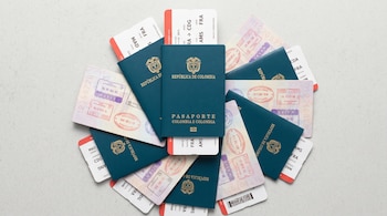 Fotografía cenital de pasaportes colombianos apilados en abanico sobre una superficie gris claro. Muestran sellos migratorios europeos y billetes de avión.