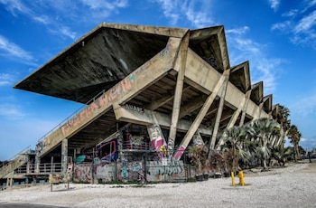 Estructura de hormigón de un estadio con tribunas escalonadas, techo voladizo, paredes con grafitis, palmeras y cielo azul con nubes