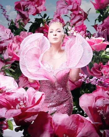 Mujer rubia con un moño alto, vestido rosa con volantes y pedrería. Rodeada de grandes flores rosas, fucsia y verdes. Fondo con cielo y nubes