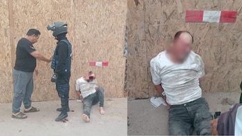 Un hombre con las manos atadas a la espalda permanece sentado en el suelo frente a un tablero de madera junto a un agente de seguridad