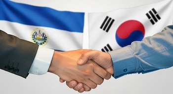 Dos manos de hombres se estrechan en el centro de la imagen. Detrás, flotan desenfocadas las banderas nacionales de El Salvador y Corea del Sur.