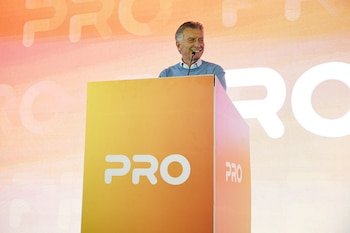 Un hombre de cabello gris sonríe detrás de un atril naranja que tiene el logo blanco de PRO. Al fondo, una pantalla con el mismo logo
