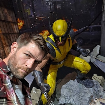 Hombre barbudo con camisa a cuadros en primer plano junto a una estatua de Wolverine con traje amarillo, negro y garras extendidas, con un fondo urbano destruido con llamas