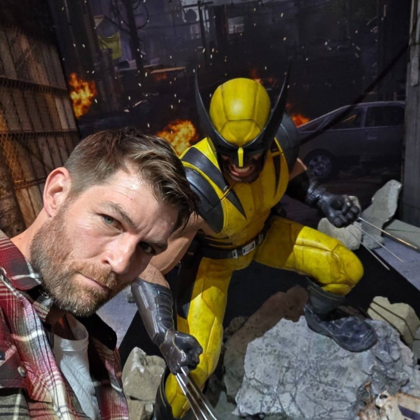 Liam McIntyre, protagonista del juego Marvel's Wolverine, posa junto a una estatua del personaje en su traje clásico, con un fondo de ciudad destruida (@liam.j.mcintyre)