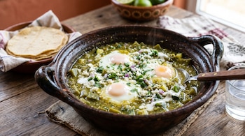 Una cazuela de barro con tres huevos en salsa verde con trozos de nopal y queso, junto a una cesta con tortillas de maíz y un vaso con agua.