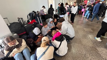 Varias personas, algunas sentadas en el suelo y otras de pie, con equipaje variado en el pasillo de una terminal de aeropuerto