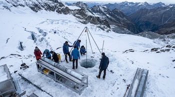 Seis científicos con equipo de perforación en un glaciar con grietas y montañas nevadas al fondo, junto a una mesa con núcleos de hielo extraídos.