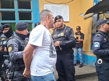 Un hombre con camiseta blanca es custodiado por cinco agentes de policía con uniformes camuflados y boinas, cerca de un edificio con ventanas