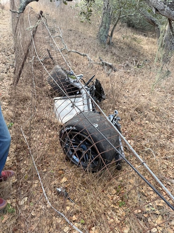 Piezas de un automóvil de carreras dañado y una rueda grande se encuentran sobre el pasto seco detrás de una red de alambre