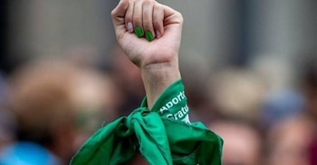 Primer plano de un puño levantado con la muñeca adornada con un pañuelo verde que dice "Aborto Legal Gratuito". Las uñas están pintadas de verde