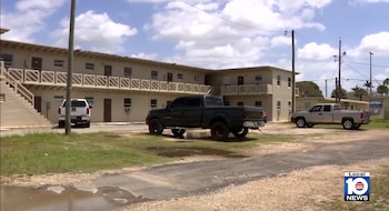 Edificio beige de dos pisos con escaleras exteriores y balcones. Tres camionetas tipo pickup estacionadas en un estacionamiento de tierra con parches de hierba. Cielo nublado