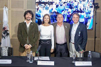 Quatro pessoas sorridentes (três homens e uma mulher) atrás de uma mesa com microfone. Ao fundo há uma bandeira com um escudo e um telão com a foto da multidão