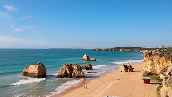 Una playa de arena con acantilados, el mar azul con olas, varias formaciones rocosas en el agua y la orilla, dos personas y una caseta de madera