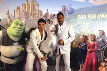 Eugenio Derbez y Eddie Murphy sostienen una figura de Burro frente a un telón con personajes y el castillo de la película Shrek 2