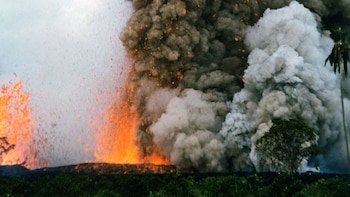 Erupción volcánica con lava naranja brillante y densa columna de humo gris que se eleva sobre vegetación oscura. Pequeñas partículas incandescentes flotan en el aire