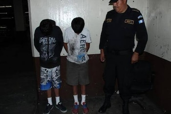 Dos jóvenes con la cabeza gacha y las manos atrás permanecen junto a un agente de policía ante una pared