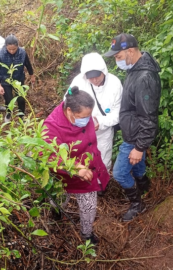 Cuatro personas, una con traje protector blanco y mascarilla, caminan por un terreno empinado con barro y ramas
