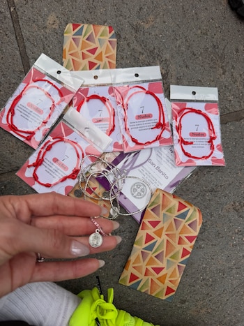 Una mano sostiene una medalla pequeña sobre varias pulseras rojas empaquetadas y sobres impresos en el suelo