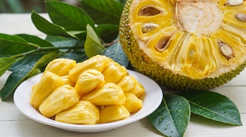 Una yaca cortada por la mitad con pulpa amarilla y semillas, porciones de yaca en un plato blanco, hojas verdes con gotas de agua y una superficie de madera blanca.