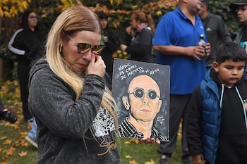 Un hombre y una mujer de mediana edad se sitúan frente a un gran estandarte rojo con el retrato de un hombre calvo con gafas de sol, iluminado por luces nocturnas