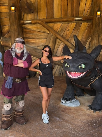 Antonela Roccuzzo sonriente posa entre un hombre disfrazado de vikingo y una gran estatua negra del dragón Chimuelo en un parque temático de madera