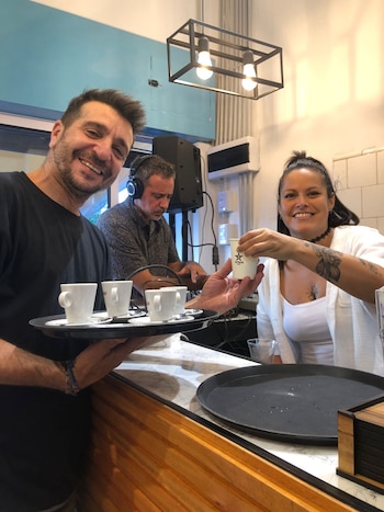 Un hombre sostiene una bandeja con tazas de café y una mujer entrega un vaso pequeño sobre un mostrador de madera
