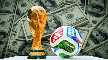 La Copa Mundial de la FIFA dorada y el balón oficial Trionda de 2026, de colores blanco, azul, rojo y verde, se muestran sobre billetes de cien dólares.