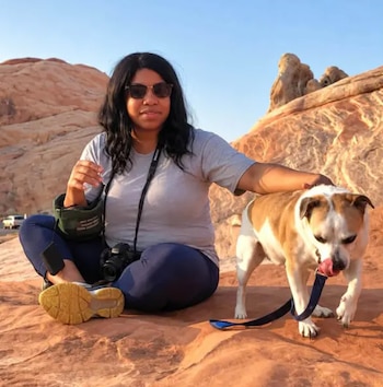 Mujer con gafas de sol y una cámara al cuello sentada en rocas de color rojizo mientras acaricia a un perro blanco y marrón