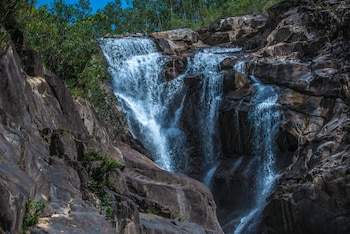 Cascada de agua cayendo sobre rocas marrones en un río rodeado por un denso bosque verde bajo un cielo azul brillante con nubes blancas