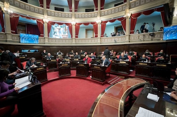 Recinto legislativo con alfombra roja, decenas de personas sentadas en escritorios de madera con sillas rojas, y un balcón superior con público. Se ven varias pantallas