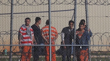 Seis hombres, algunos vistiendo uniformes naranjas de detenido, se encuentran detrás de una cerca de malla ciclónica con alambre de púas, bajo un cielo soleado