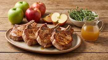 Cuatro costeletas de cerdo en una fuente, junto a manzanas verdes y rojas, una jarra con caldo y un cuenco con hierbas sobre una mesa.