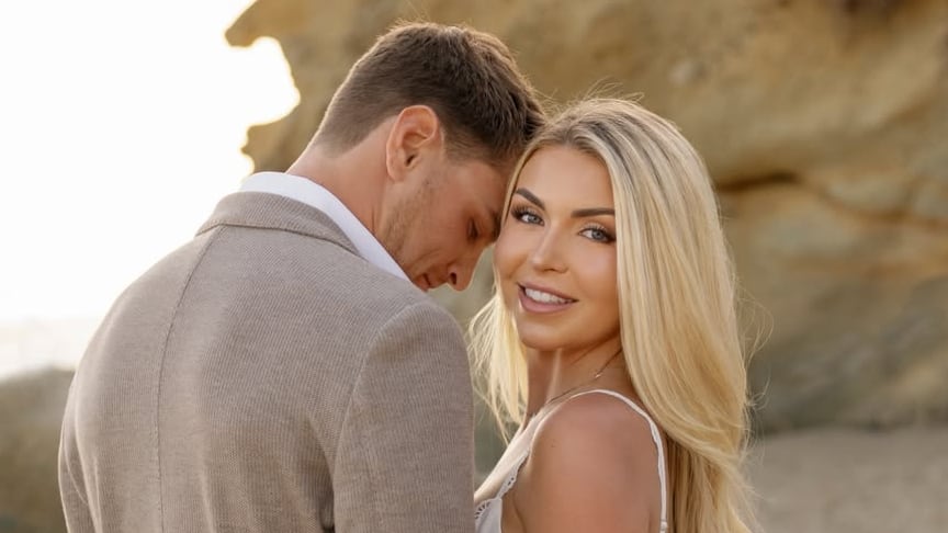 El expiloto de F1 Logan Sargeant y su pareja posan tomados de la mano en una playa de arena, celebrando su compromiso.