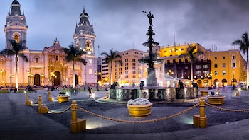 Vista panorámica de una plaza iluminada al atardecer con una fuente central, una catedral con dos campanarios y edificios históricos alrededor, palmeras y personas